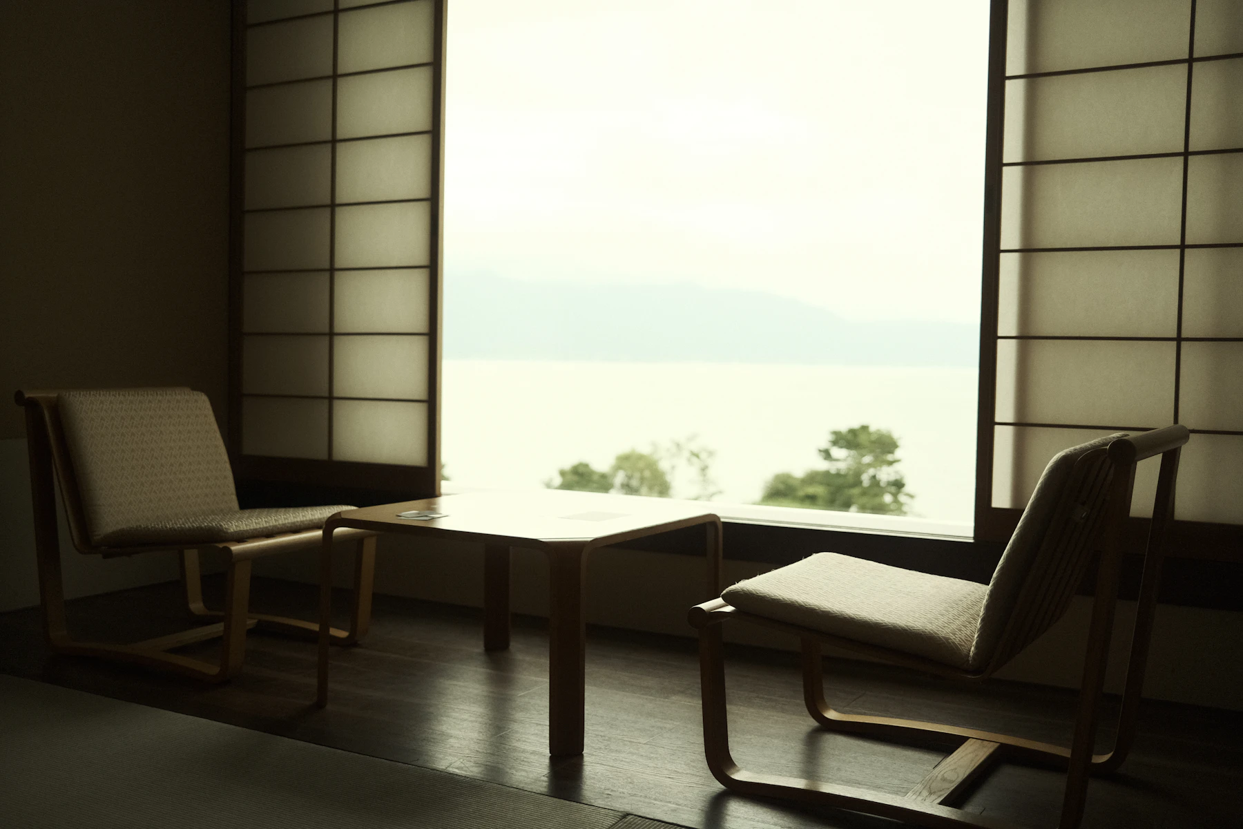 障子のある客室の窓辺に置かれた椅子と座卓、曇り空の湖